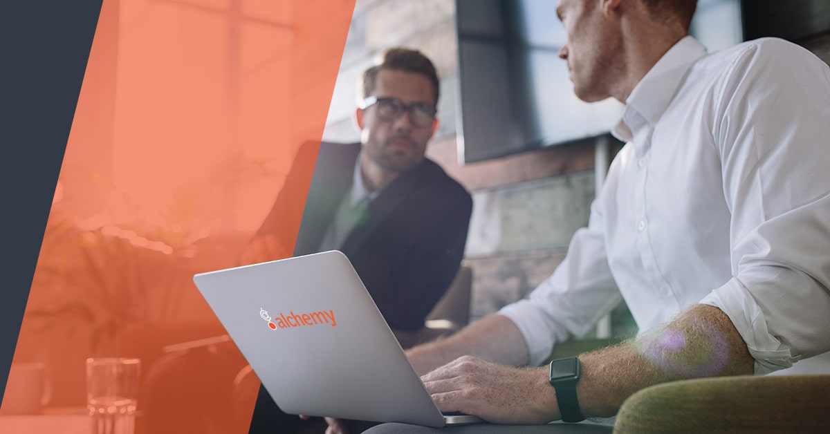 Two professionals are having a discussion in an office setting, one using a laptop with an Alchemy logo on it.